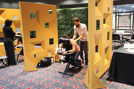 Masseuse giving a chair massage in front of a green shrub wall.