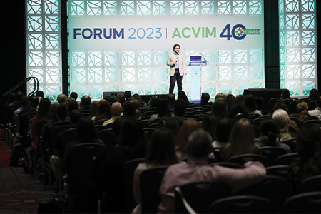 Man speaking in front of dark audience with the ACVIM Forum logo behind him.
