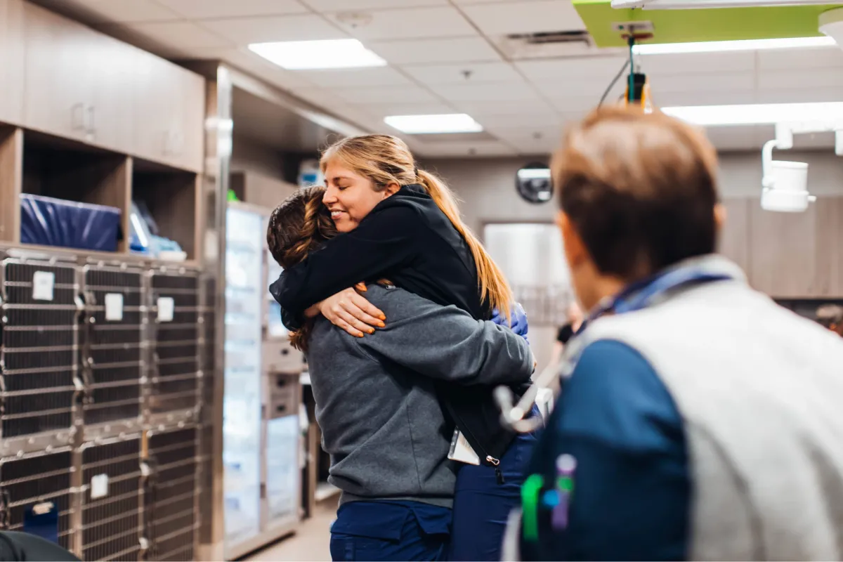 Two veterinary professionals smile and embrace in an exam room
